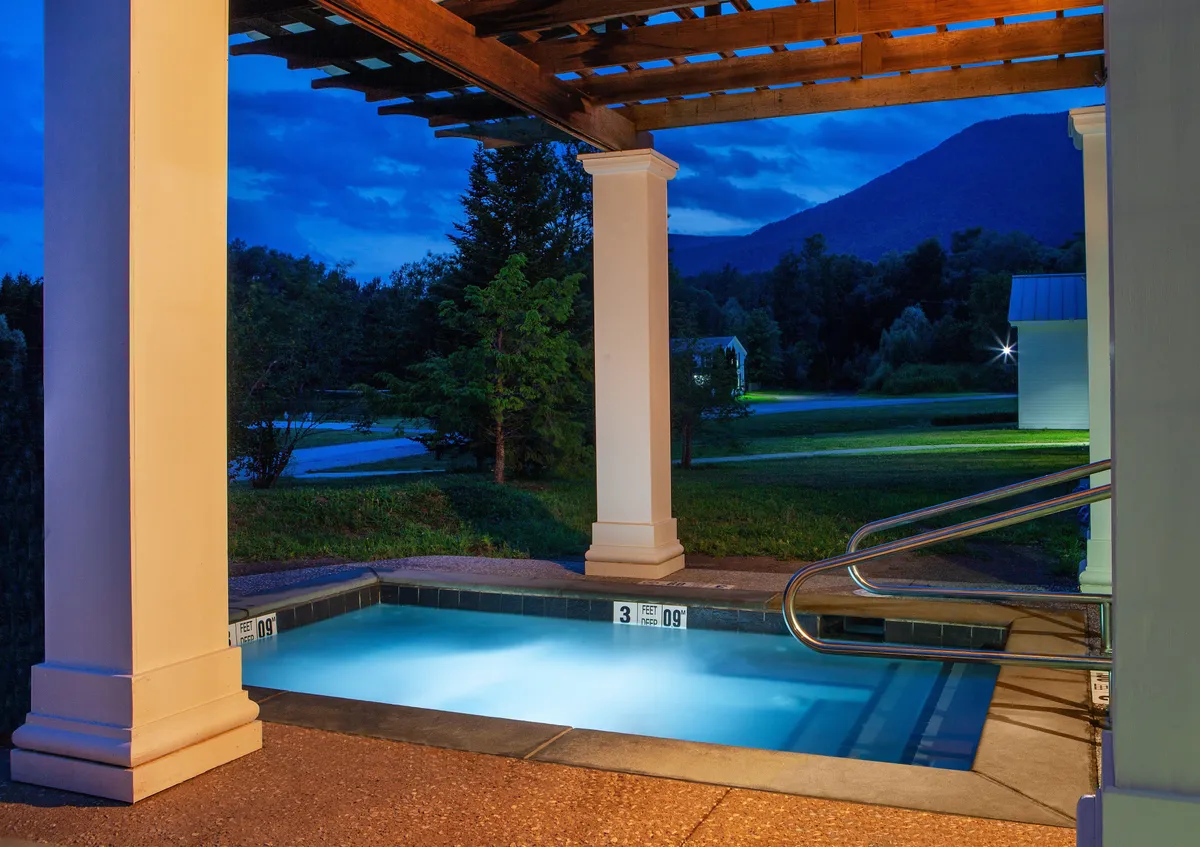 A heated swimming pool glows invitingly under the evening sky, sheltered by a wooden pergola and surrounded by lush trees with views of distant mountains. The tranquil setting is perfect for evening swims and stargazing, evoking a sense of serenity and intimacy.