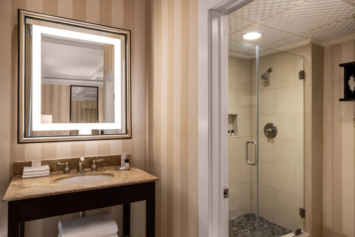This image presents a spa-inspired bathroom with a well-lit vanity mirror, modern sink area, and a glass walk-in shower enclosure bathed in gentle morning light. The space feels serene and inviting, emphasizing relaxation and refreshment.