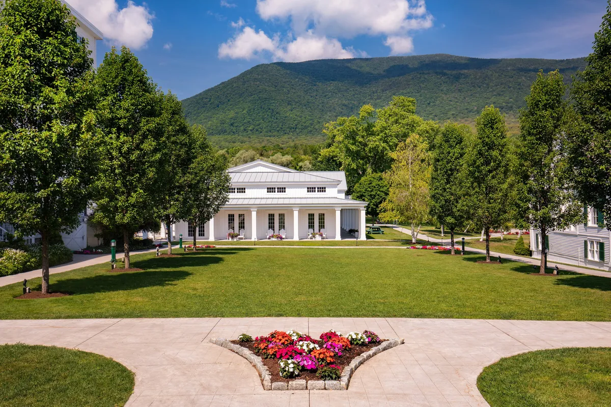 A beautifully manicured lawn bordered by vibrant flower beds unfolds before the classic architecture of The Equinox Resort, with mountains rising in the background. The soft light of sunset creates a warm, welcoming ambiance, perfect for outdoor activities or a peaceful stroll.