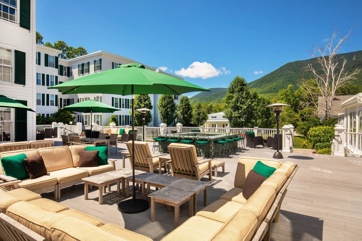 An inviting outdoor dining terrace with green umbrellas, comfortable sofa seating, and tables set against a stunning mountain backdrop. The scene depicts guests enjoying relaxed meals, drinks, and conversation, with a leisurely, celebratory mood at sunset.