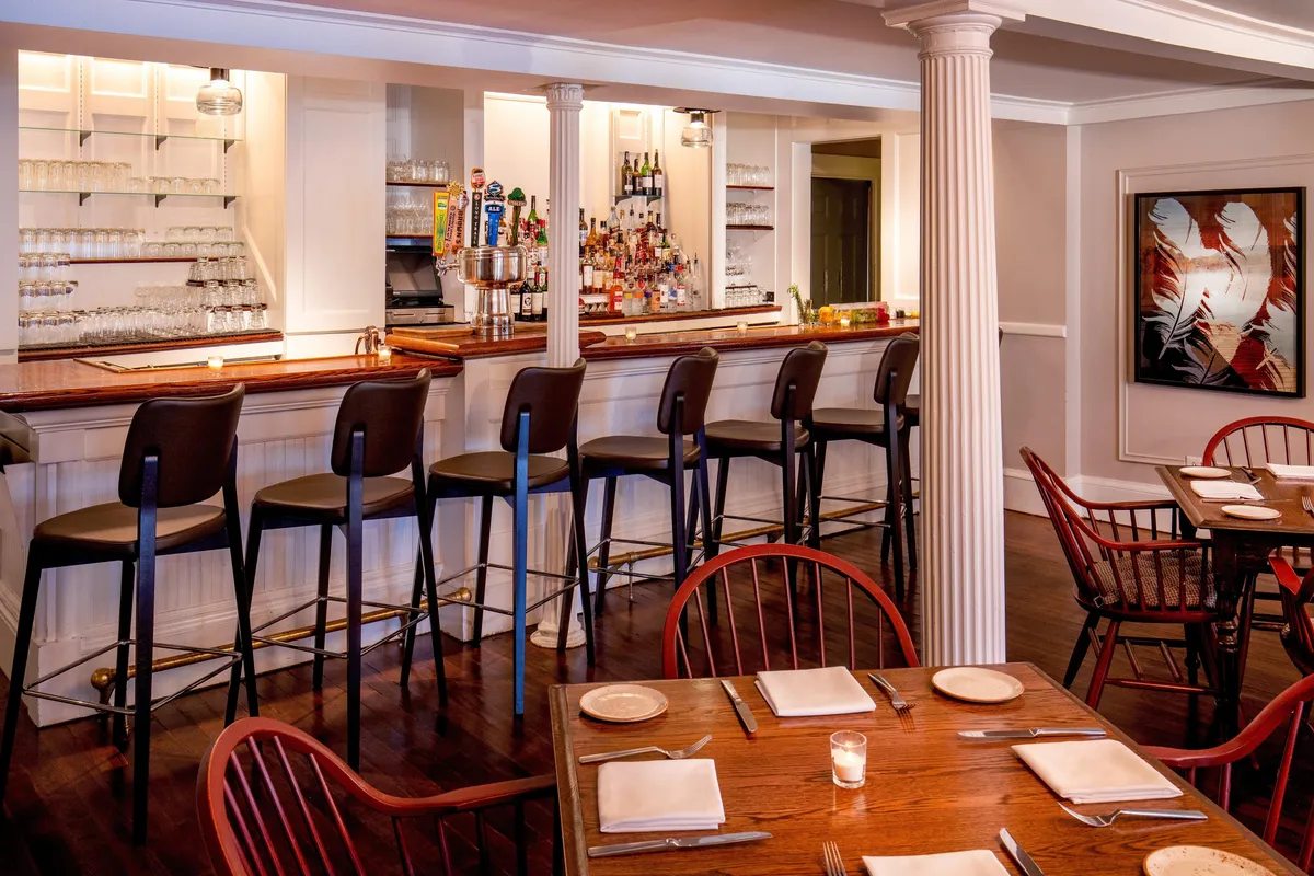 A stylish interior dining and bar space with a modern bar area, surrounding dining tables, and a striking piece of art on the wall. The area is softly lit, creating a welcoming environment ideal for socializing, enjoying meals, or savoring drinks—a hub of guest activity and convenience.