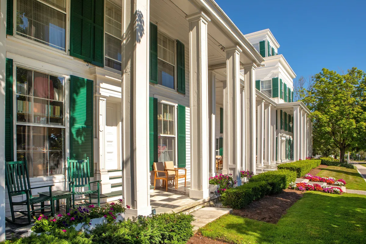 A classic front porch featuring green shutters, white columns, rows of rocking chairs, and blooming flower beds. The scene invites guests to unwind with morning coffee or evening drinks, offering a welcoming, communal vibe.