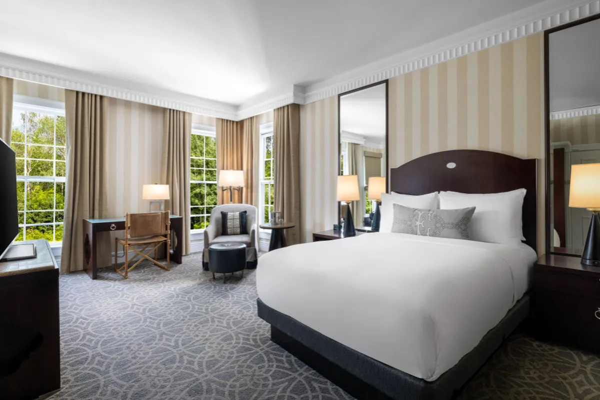 The image showcases a serene studio interior at The Equinox Golf Resort & Spa. A plush bed with crisp white linens sits near large windows that fill the space with natural light. Nearby, an inviting armchair and a sleek desk offer cozy spots for relaxation or work. Elegant mirrors add depth, and the overall atmosphere is tranquil and welcoming.