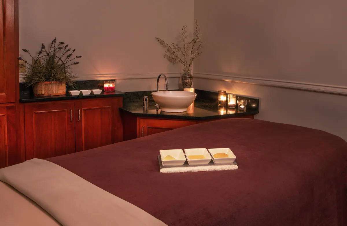 The image shows a tranquil interior spa room featuring a massage table, ceramic bowls, a sink, candles, and a decorative plant. The soothing setup is designed for relaxation and self-care, with soft lighting and thoughtful decor promoting a serene ambiance.