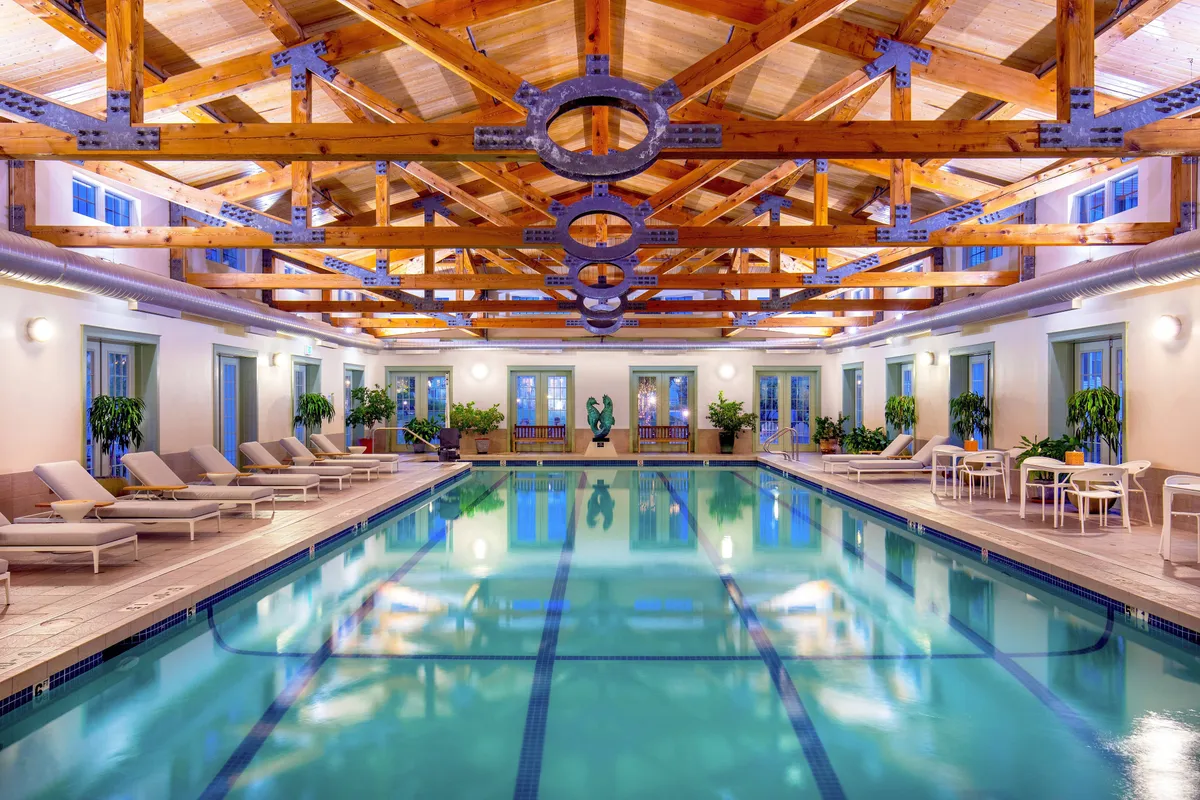 A luxurious indoor swimming pool area featuring wooden beams overhead, elegant lounge chairs neatly arranged beside the water, and carefully curated local artwork and lush plants adding a tranquil and sophisticated ambiance. The scene exudes a sense of comfort and exclusivity, inviting guests to relax and unwind in a serene environment.
